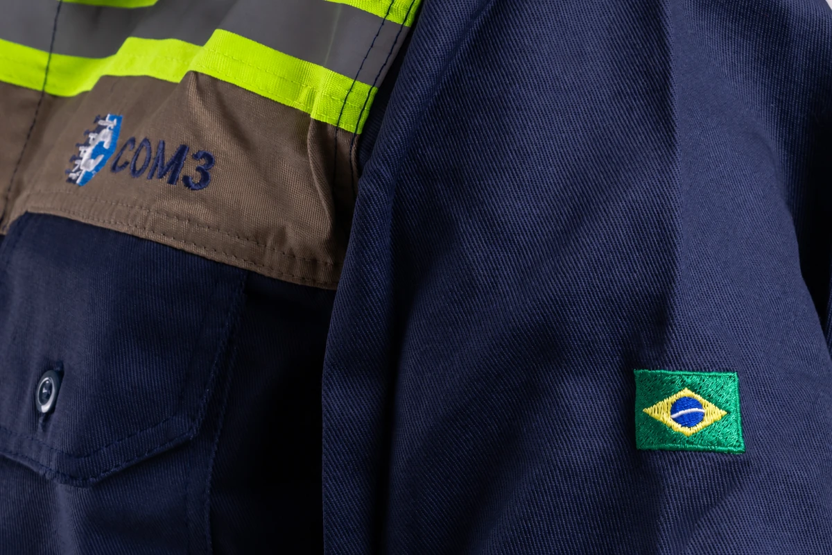 Detalhe do jaleco azul da COM3 com a logo COM3 e a bandeira do Brasil, em foto editada por IA.